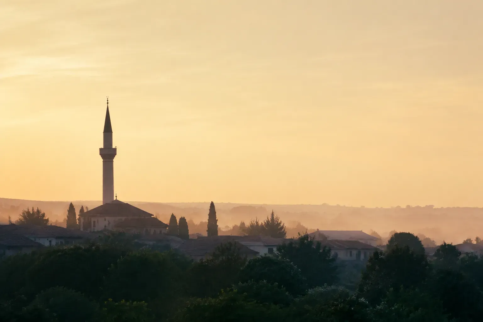 Minaret et toits d'une ville au lever du jour, ciel doré et arbres en contrebas